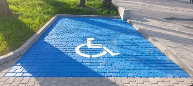 Parking dla niepełnosprawnych z niebieskiej kostki brukowej, biały symbol wózka inwalidzkiego. Miejsce parkingowe wydzielone dla osób z ograniczoną sprawnością ruchową, idealne do oznaczeń infrastruktury miejskiej, budowlanej i oznakowania parkingów