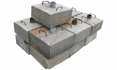 Stos prostokątnych, betonowych podstaw do znaków drogowych na białym tle. Każdy blok wyposażony jest w dwa metalowe uchwyty do przenoszenia oraz centralnie umieszczoną metalową tuleję na słupek