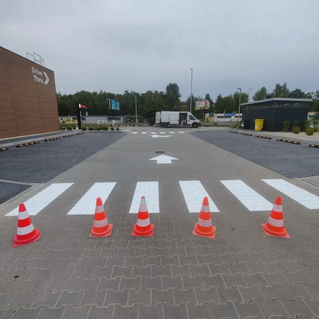 Świeżo wymalowane białe pasy przejścia dla pieszych i strzałki kierunkowe na parkingu, zabezpieczone pachołkami